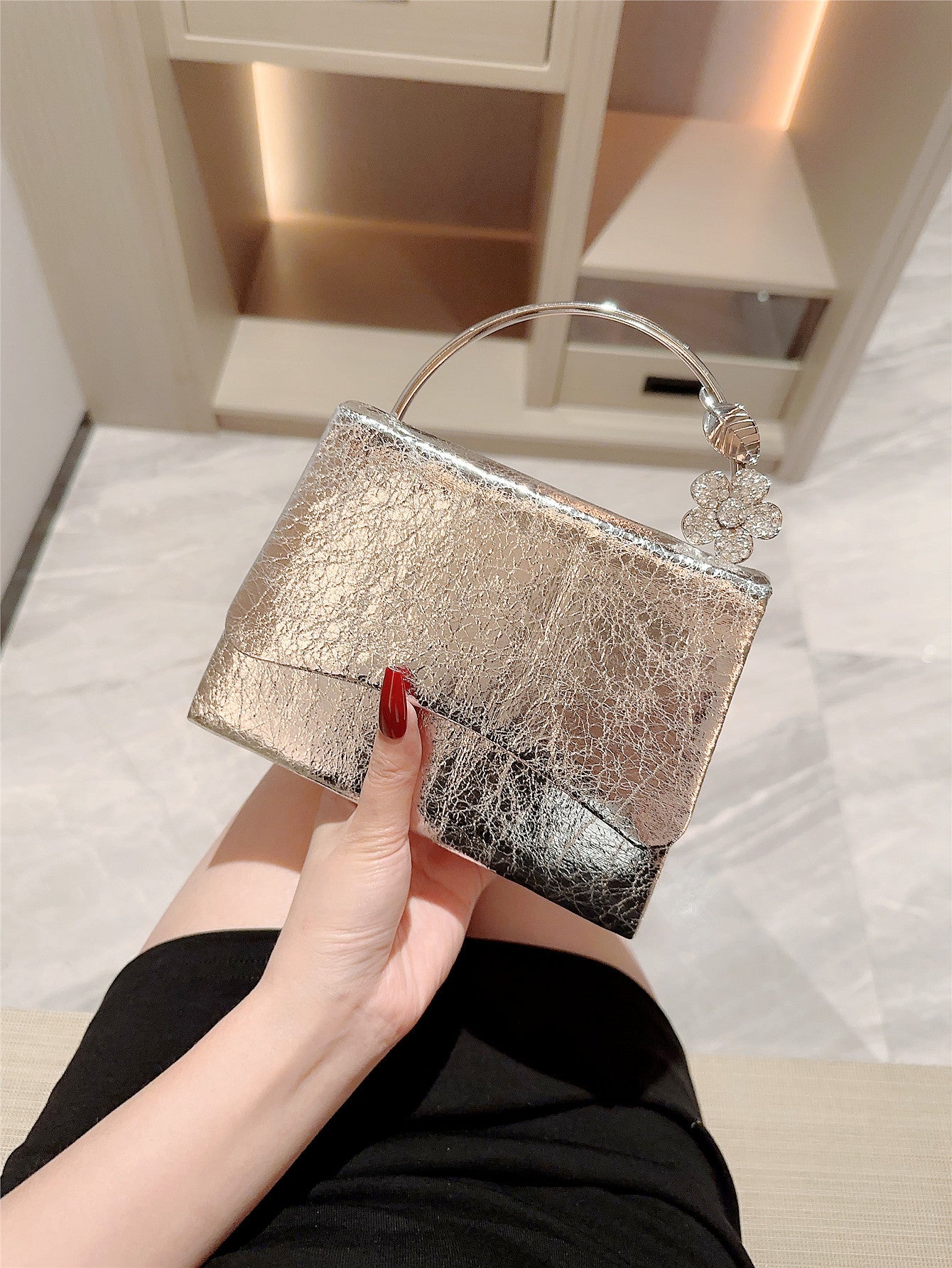 Hand holding a silver clutch bag with a decorative handle that has a flower on a light-colored floor.
