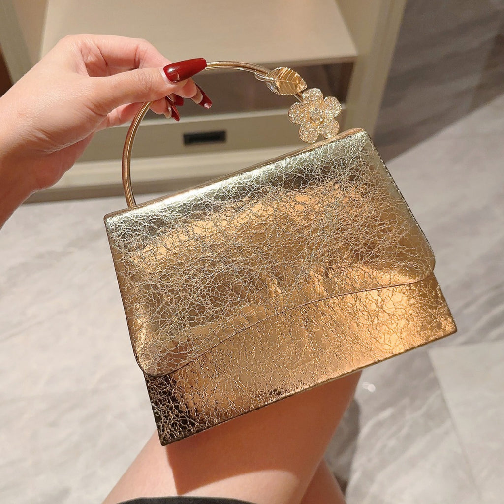 Gold clutch bag with floral handle held by a hand against a marble background