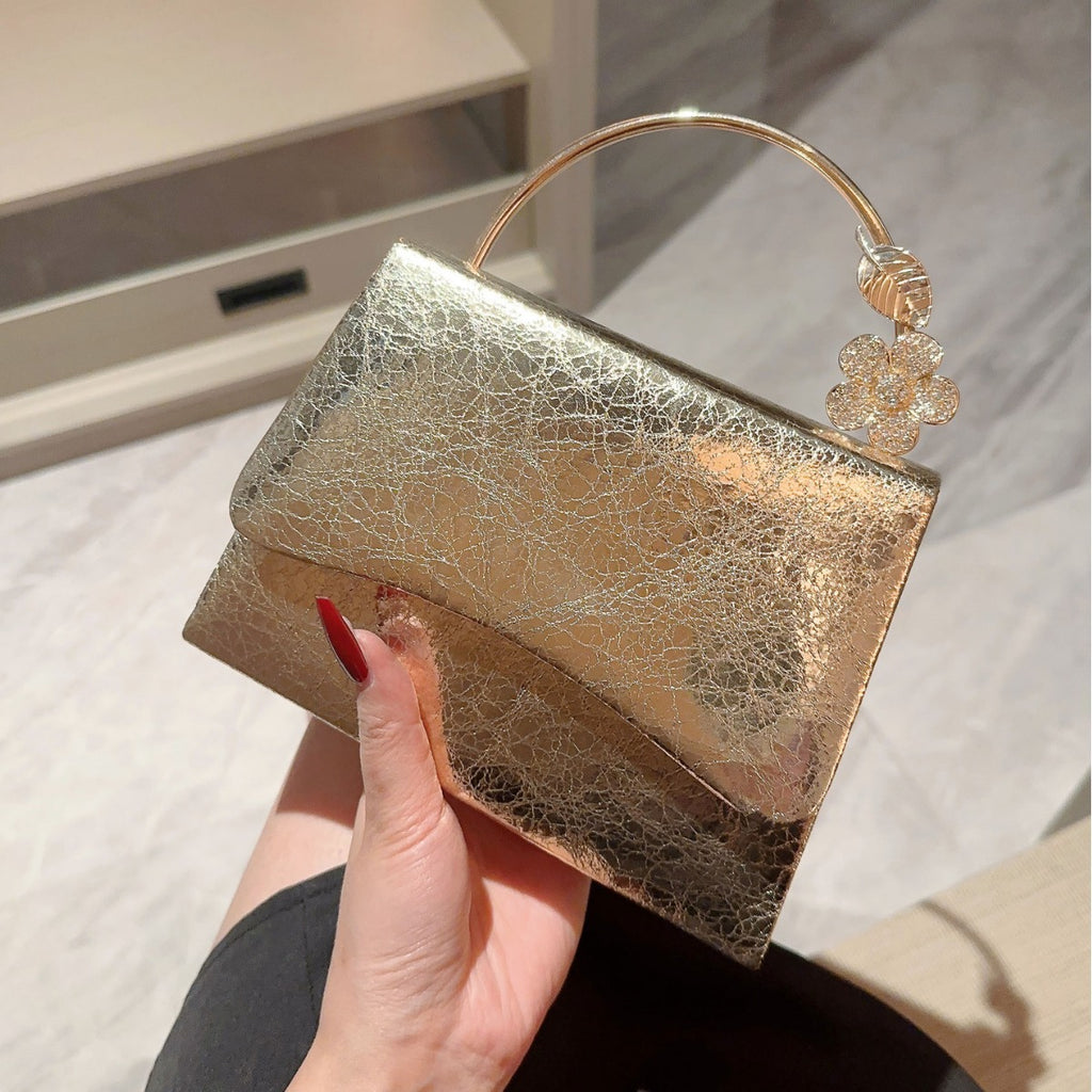 Gold clutch bag with decorative handle held by a hand against a neutral background