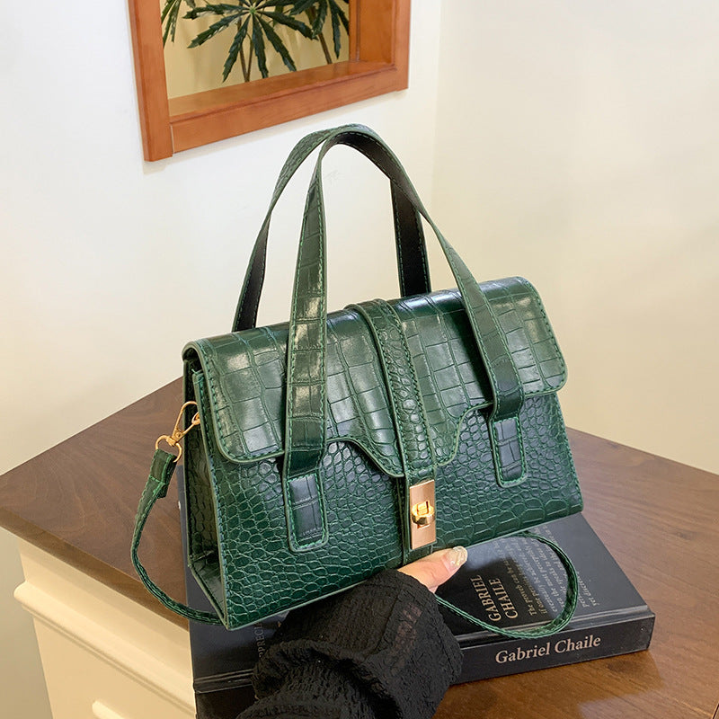 Green crocodile-patterned handbag held by a person on a wooden surface with a book underneath.